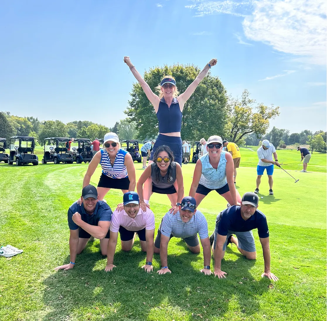 Eight golfers stacked in a human pyramid on the fairway, the one on top with both arms raised