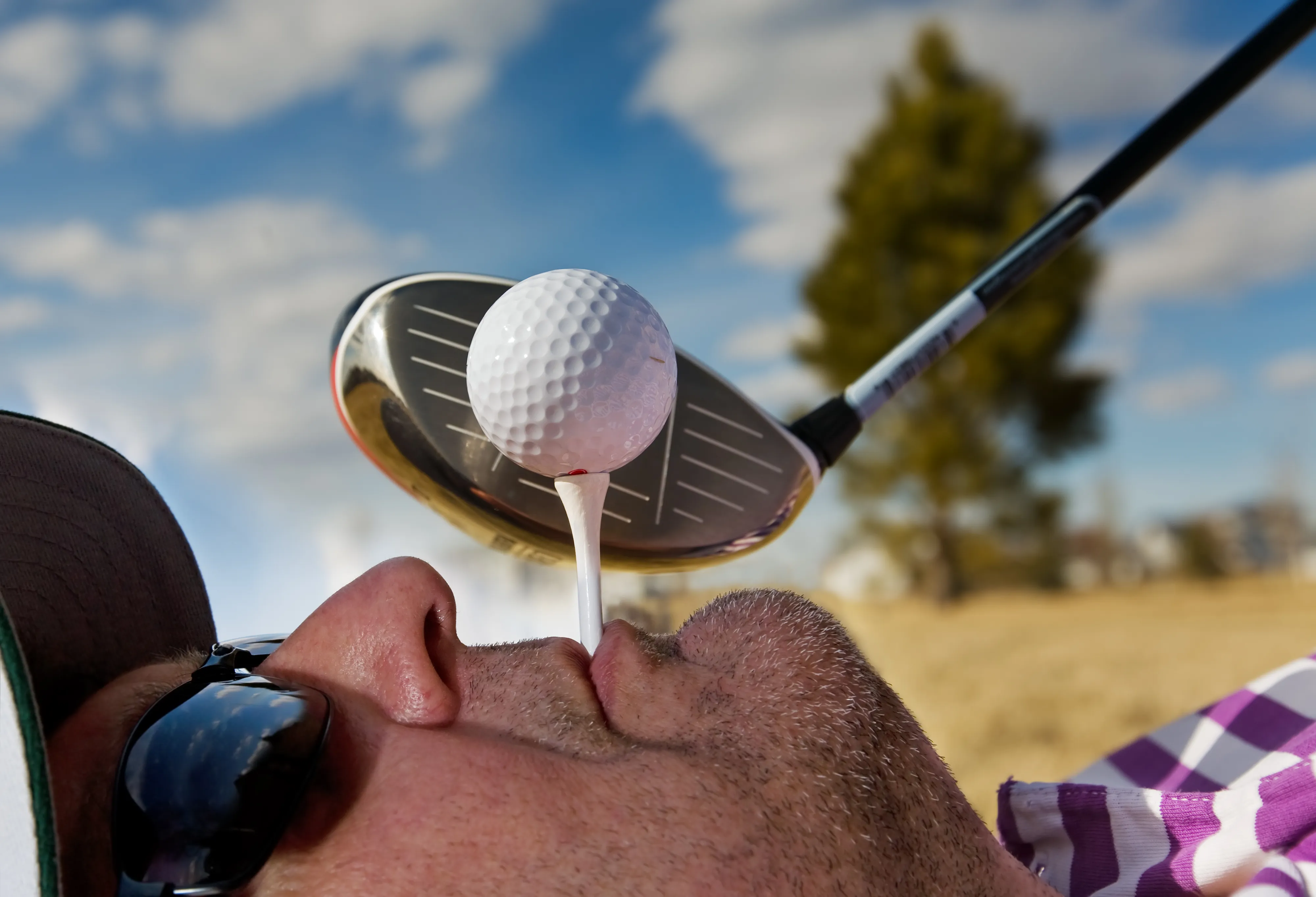 A close-up of a golfer balancing a teed-up ball at his mouth, driver poised above it