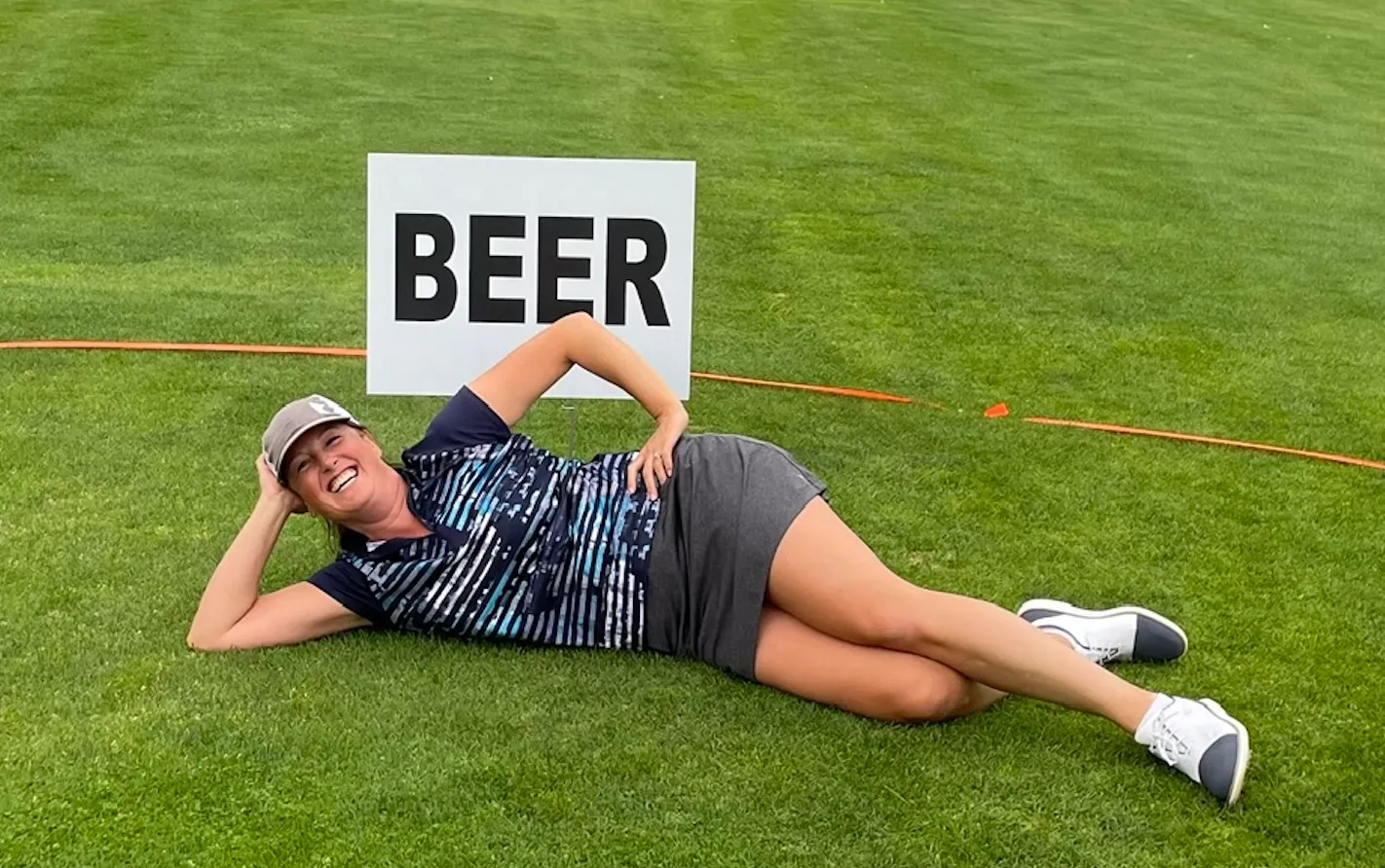 A golfer laughing on the fairway beside a hole sign that reads BEER